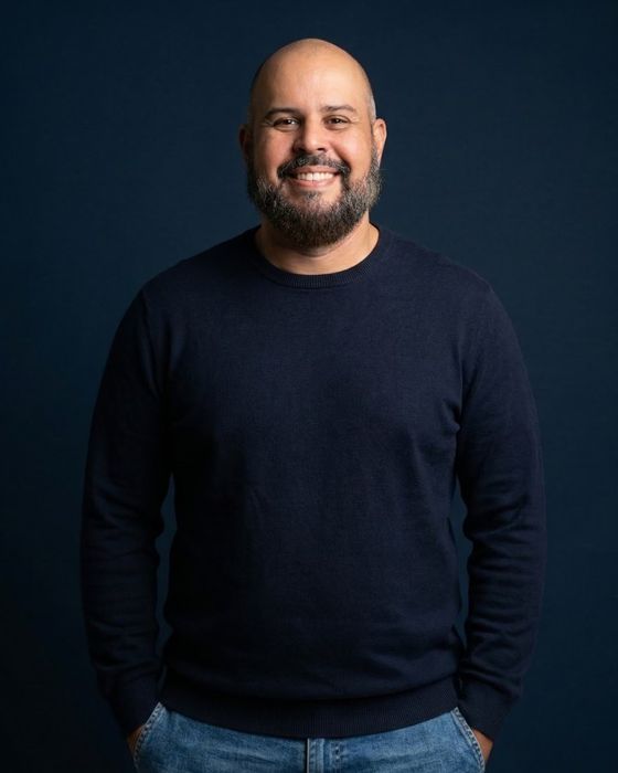 Fábio Carigé sorrindo, de suéter azul-marinho, em fundo azul escuro